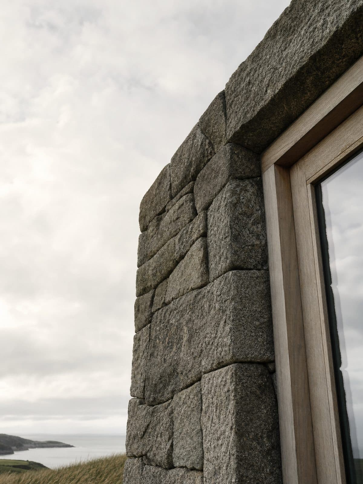 A granite spine wall meeting an oak-framed opening, photographed against a bright coastal sky.