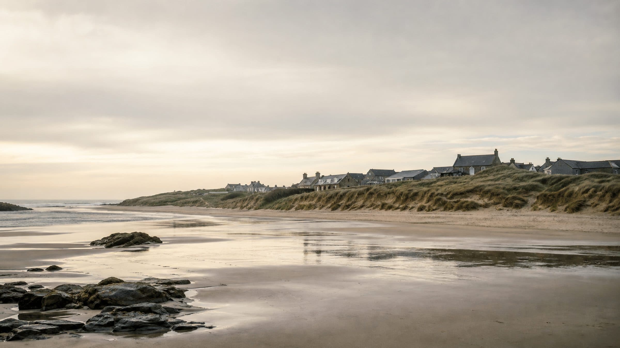 A wide south-facing sand bay at low tide, with protected dunes and a low village roofline behind them.
