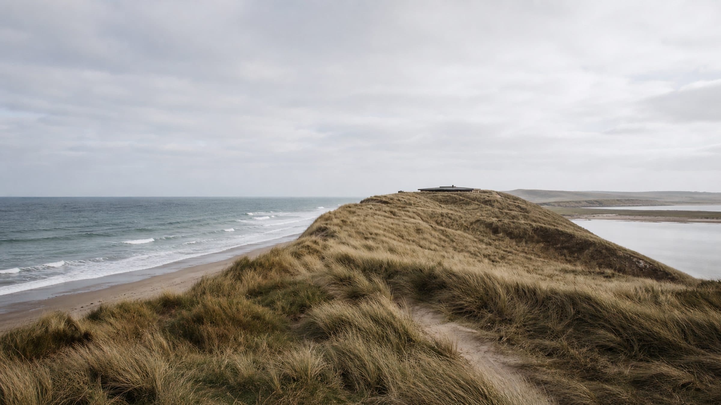 A low dune peninsula reaching into the Atlantic, held by marram grass, with a single low roofline just visible.
