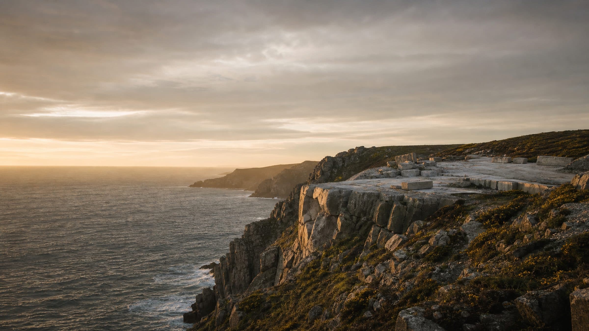 A granite headland falling to the Atlantic in low evening light, with a level shelf of cut stone near the cliff edge.