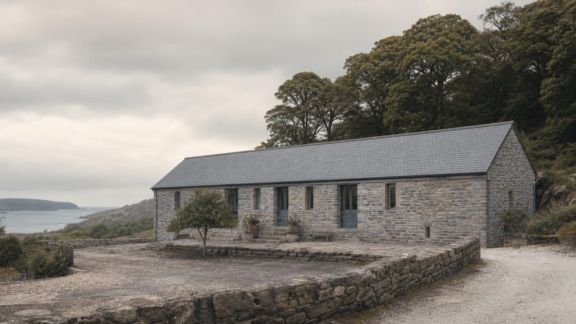 A long stone barn with a new oak roof standing in a walled yard, oak woodland rising behind it.