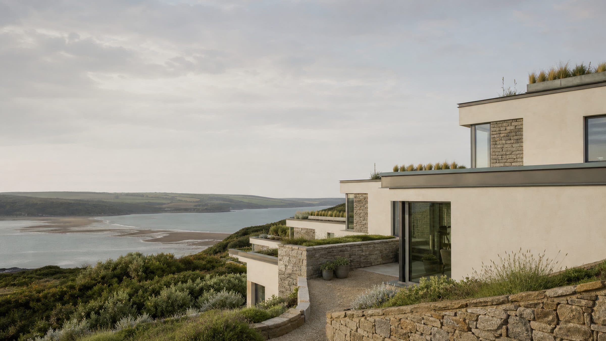 A stepped courtyard house on a sheltered hillside, rendered walls and granite facing, with an estuary beyond.