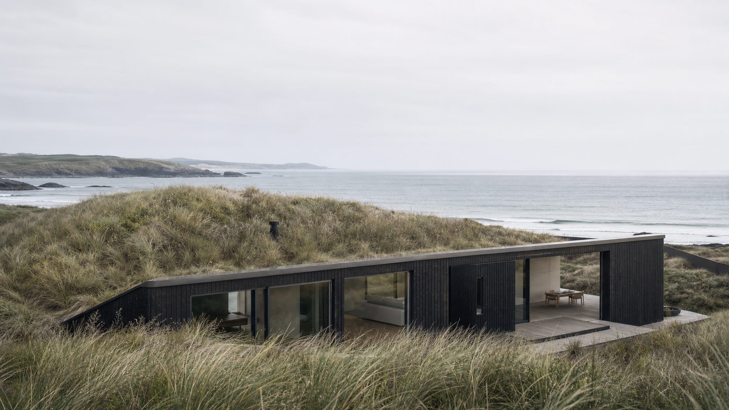 A low char-treated timber house set into a dune, its roof below the dune crest, with the Atlantic behind.
