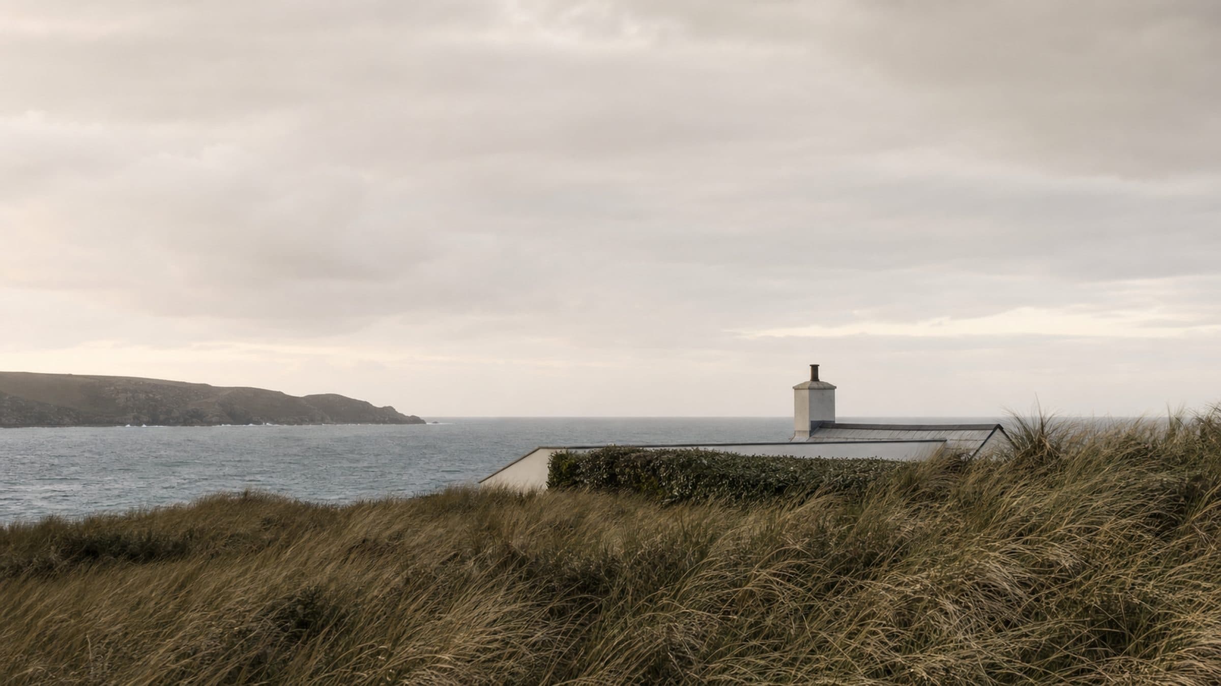 A lime-rendered courtyard house set low behind dunes, its chimney and hedge line visible from the bay.
